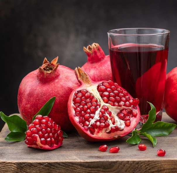 Image shows 3 harvested pomegranate Provence fruits on a wooden chopping board beside a glass of pomegranate juice. One of the fruits is halved, showing its red seeds. There are 4 seeds plus some green leaves on the board.
